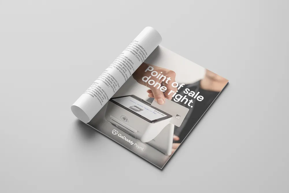 A4 print ad on a gray surface with its top sheet rolled back into a tube, revealing the page beneath: a hand inserting a chip card into a white Poynt terminal, under the headline 'Point of sale done right.' and the GoDaddy Poynt lockup.