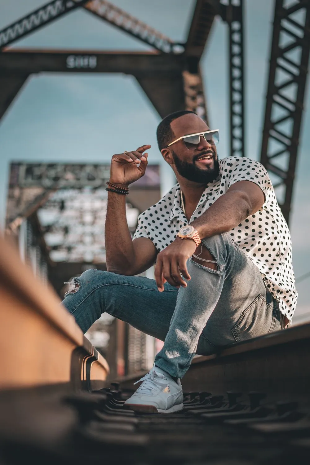 Man in a white polka-dot shirt, ripped jeans and white sneakers seated on the rails of a steel truss railway bridge, shot from a low angle against a pale blue sky.