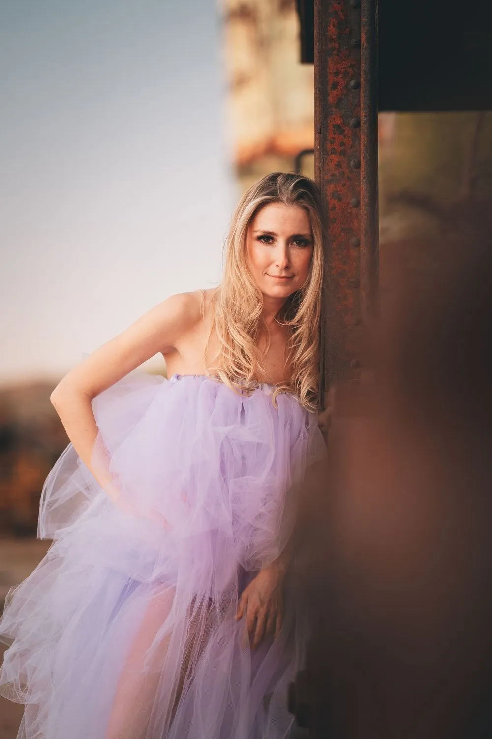 Woman in a voluminous lilac tulle gown leaning against a rusted steel girder, one hand on her hip, warm open sky behind her.