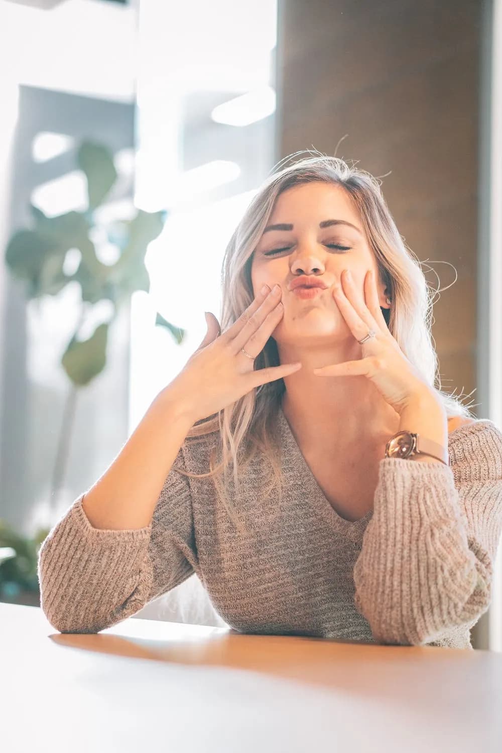 Woman in a knit sweater at a white table, eyes closed and hands framing her face, backlit by a bright window with a houseplant behind her.