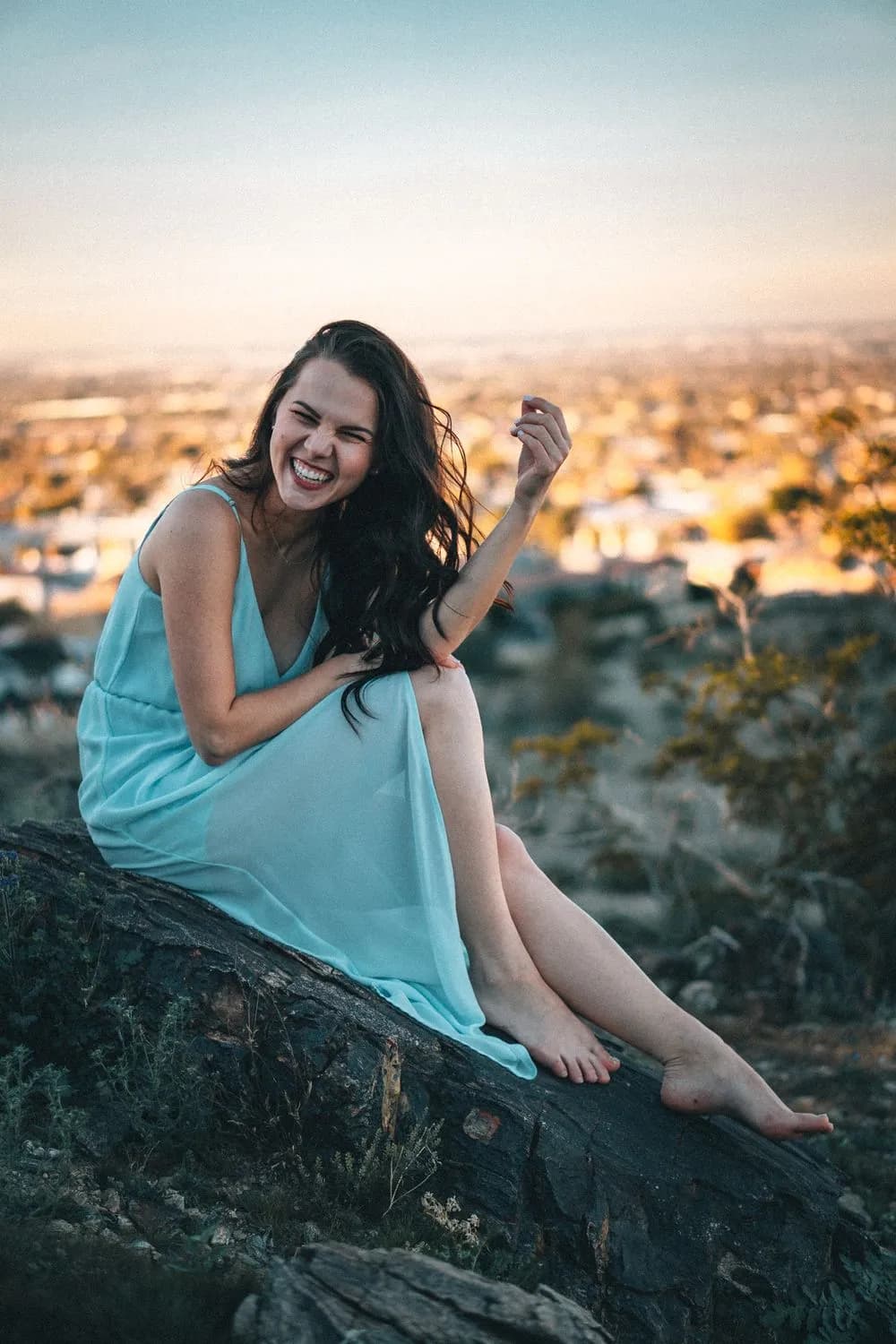 Woman in a pale turquoise slip dress sitting barefoot on a dark rock outcrop, laughing, with a city skyline glowing gold behind her at dusk.
