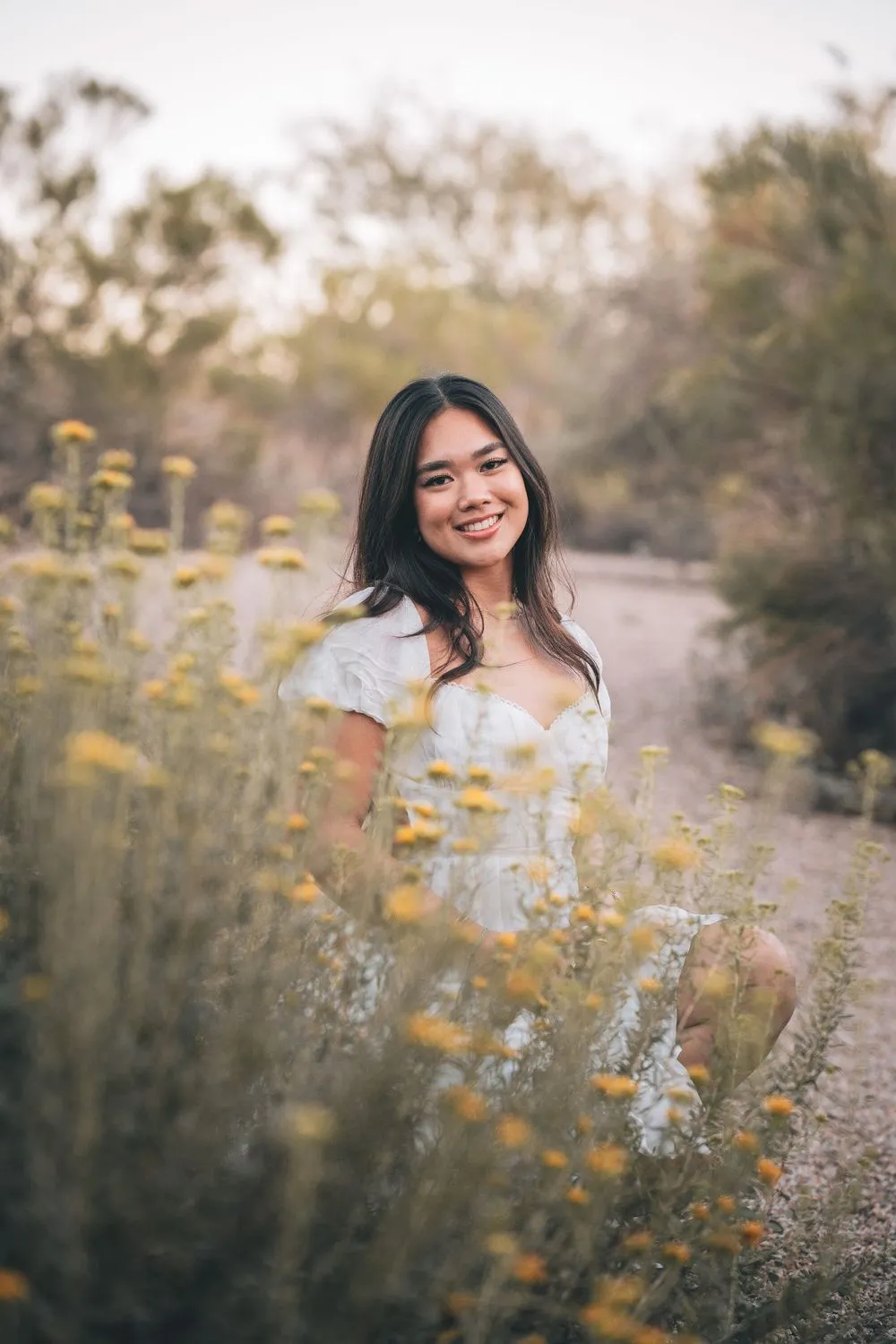 Woman in a white puff-sleeve dress crouching among tall yellow desert wildflowers, smiling at the camera, with desert scrub soft behind her.