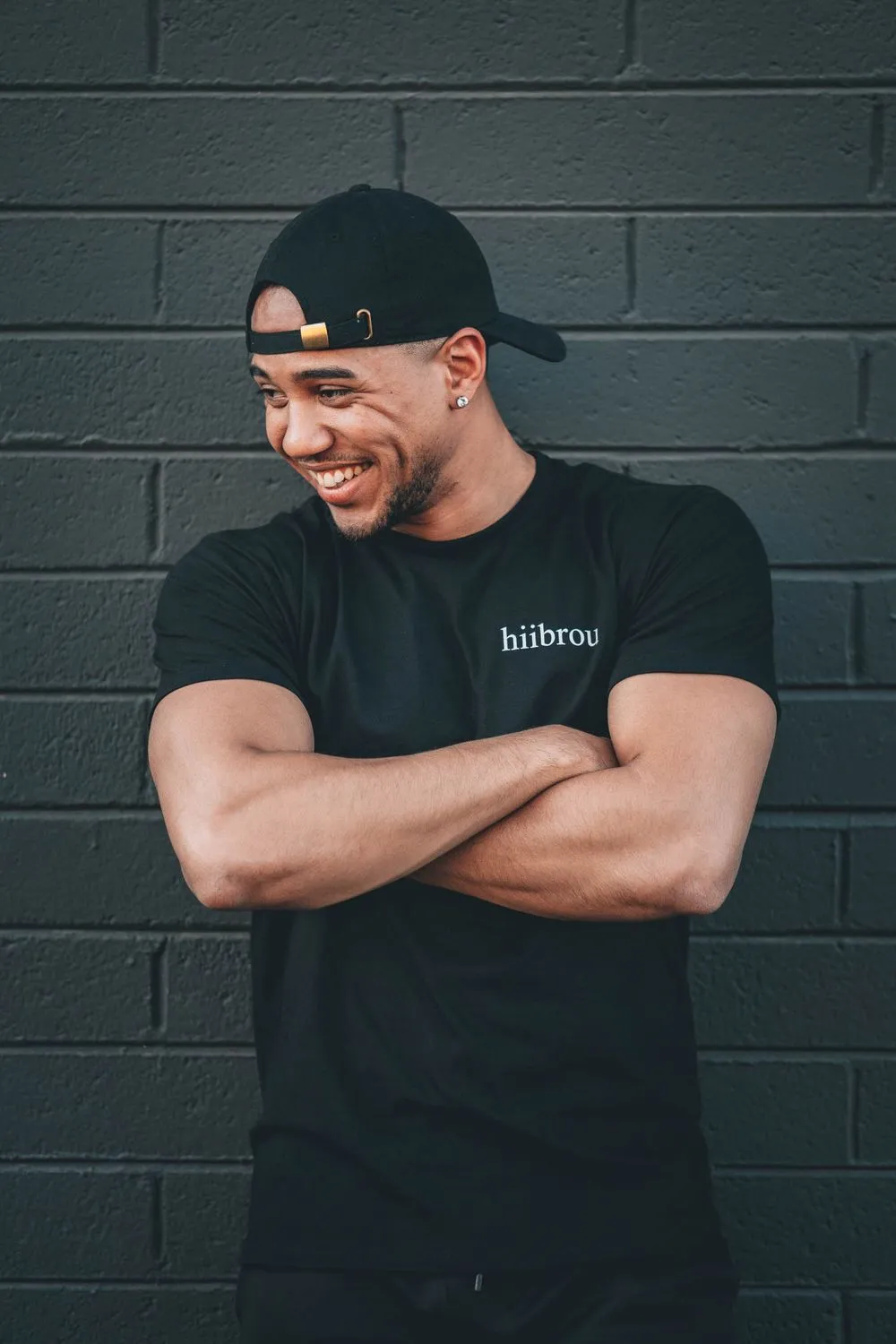 Man in a black printed t-shirt and a backwards black cap, arms crossed and laughing off-camera, against a dark gray painted brick wall.