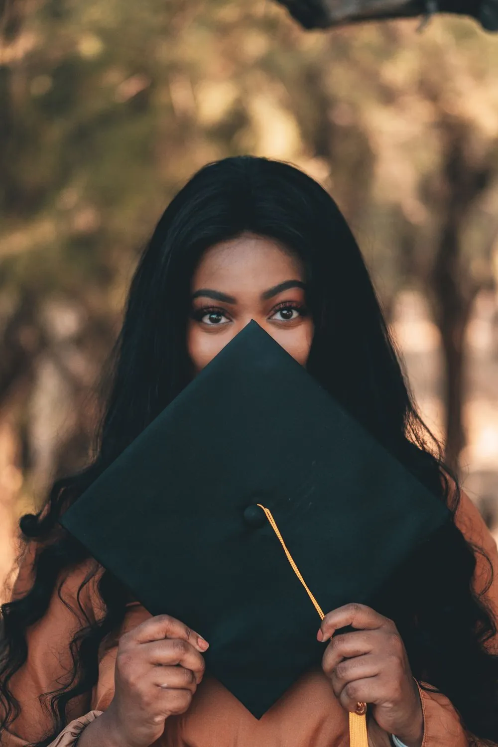 Woman holding a black graduation cap with a gold tassel across the lower half of her face, eyes level with the camera, warm out-of-focus trees behind her.