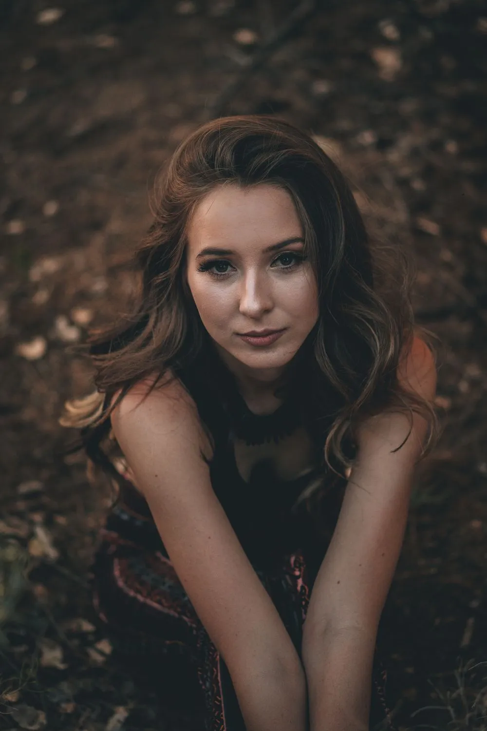 Woman with long wavy hair seated on dark forest ground in a patterned dress, arms folded across her knees, looking up into the camera in warm low light.