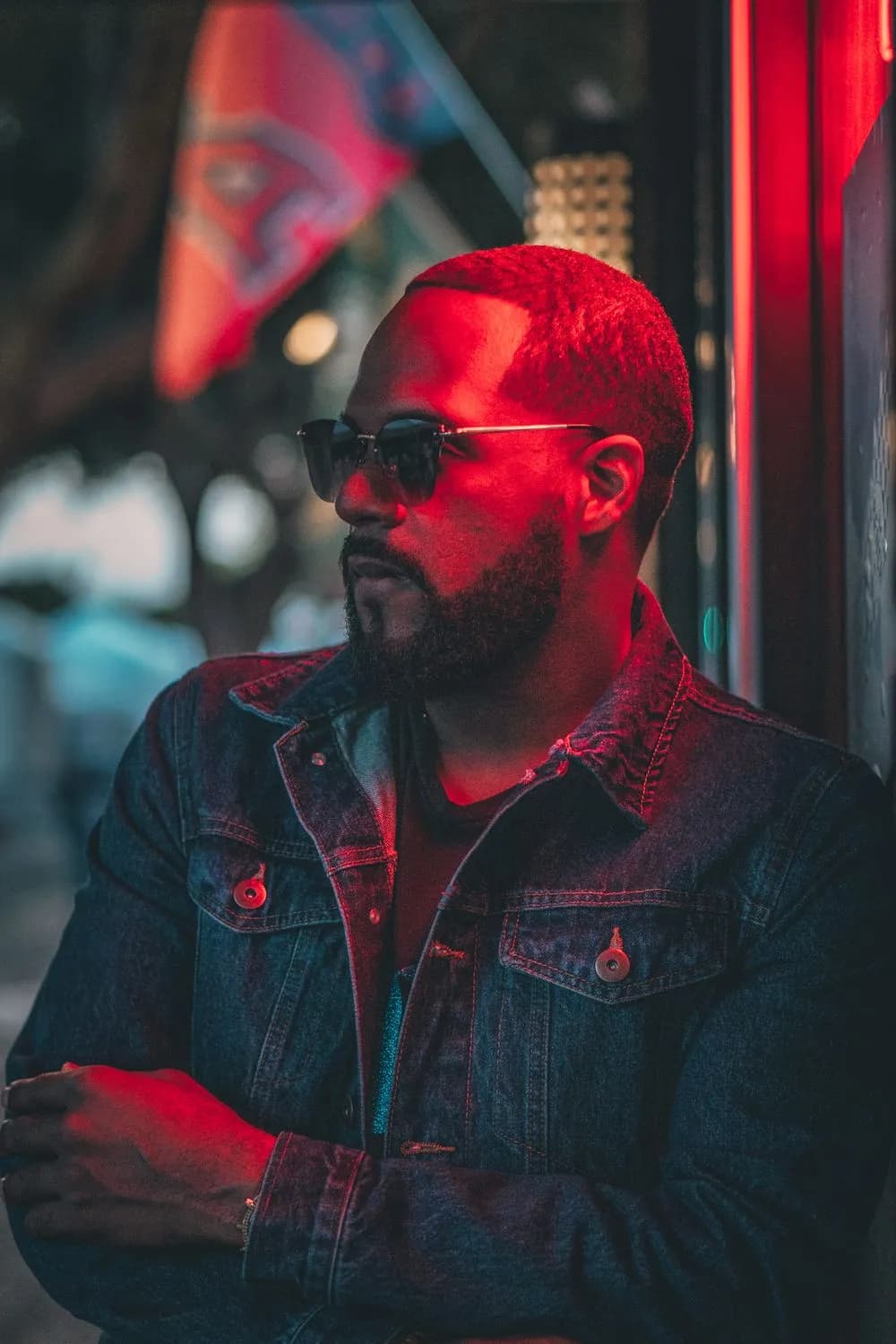 Bearded man in dark rectangular sunglasses and a denim jacket, arms folded, leaning against a storefront and washed in hard red neon light, with blurred signage glowing behind him.